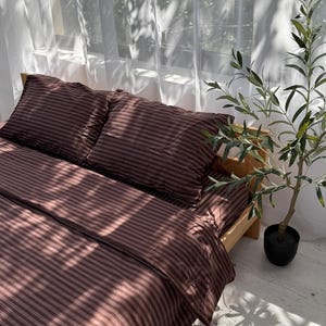 Op de afbeelding: Een bed met een bruin gestreept dekbedovertrek en kussenslopen. Het bed staat op een houten frame, met een kleine plant in een pot ernaast. De kamer heeft een raam met transparante witte gordijnen.