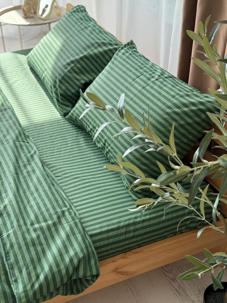 a bed with green and white striped bedding, including pillows and a blanket, placed on a wooden bed frame. There is a plant in the foreground, adding a touch of greenery to the scene.