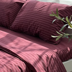 May include: A burgundy striped bedding set, including a fitted sheet and two pillowcases, is neatly arranged on a white bed frame. The fabric has a soft, textured appearance. Natural light streams in, casting shadows and highlighting the details.