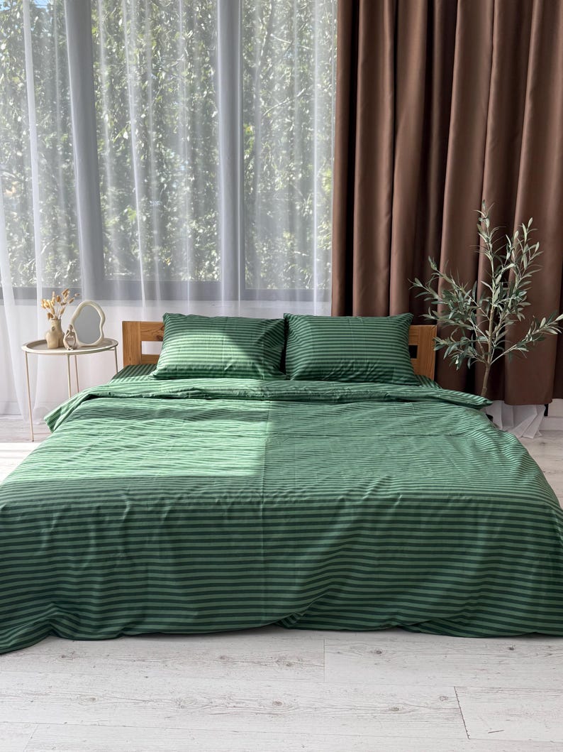 a bedroom with a large bed covered in a green and white striped comforter. The bed is positioned against a window with brown curtains, and there is a small table with a vase of flowers on it.