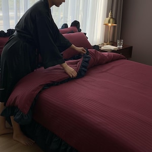 A person in a black robe is making the bed with a red and black striped comforter and matching pillows.