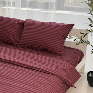May include: Burgundy striped bedding set, including a flat sheet, fitted sheet, and two pillowcases. The sheets have a subtle, textured appearance and are neatly arranged on a white bed frame. The room is bright with natural light.