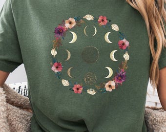 May include: Olive green t-shirt featuring a floral and moon phase design. The graphic includes gold crescent moons, a central full moon, and a wreath of pink and purple flowers with gold leaves. The shirt has a crew neck and short sleeves.