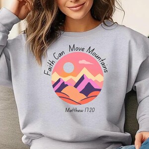 May include: A pale grey sweatshirt with a circular graphic depicting a mountain range in pinks, oranges, and purples, with a sun and cloud. The text "Faith Can Move Mountains" and "Matthew 17:20" are printed on the sweatshirt.