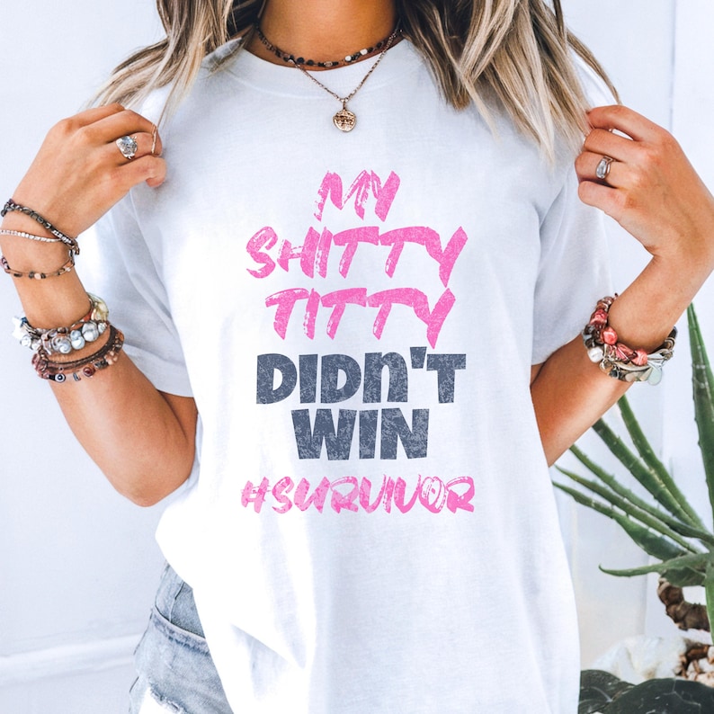 May include: White t-shirt with the text "MY SHITTY TITTY DIDN'T WIN #SURVIVOR" in pink and gray lettering. The shirt is being worn by a person with blonde hair and multiple bracelets.