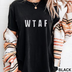 K&ouml;nnte beinhalten: Schwarzes T-Shirt mit den wei&szlig;en Buchstaben "WTAF" auf der Vorderseite. Das Shirt wird mit einem gemusterten Pullover getragen. Das Wort "BLACK" ist in der unteren rechten Ecke sichtbar.