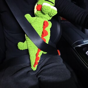 K&ouml;nnte beinhalten: Ein gr&uuml;nes, geh&auml;keltes Alligator-Spielzeug ist mit einem schwarzen Sicherheitsgurt in einem Auto befestigt. Der Alligator hat rote und wei&szlig;e Details.