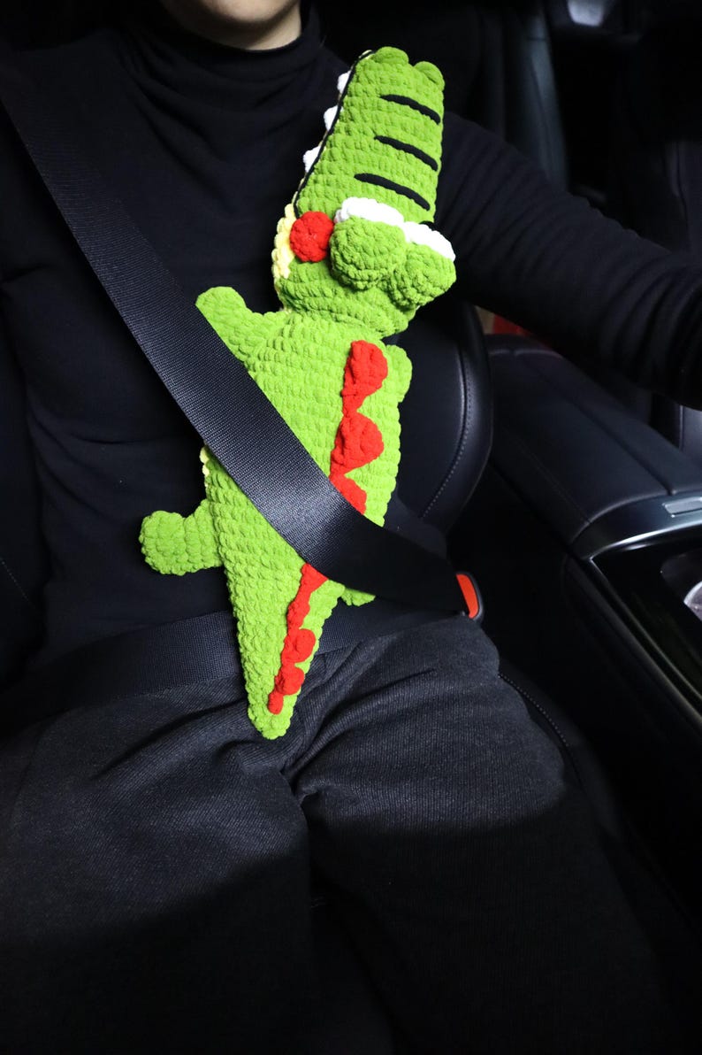 K&ouml;nnte beinhalten: Ein gr&uuml;nes, geh&auml;keltes Alligator-Spielzeug ist mit einem schwarzen Sicherheitsgurt in einem Auto befestigt. Der Alligator hat rote und wei&szlig;e Details.