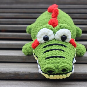 K&ouml;nnte beinhalten: Gr&uuml;nes, geh&auml;keltes Alligator-Pl&uuml;schtier mit roten und wei&szlig;en Augen und einem roten, noppigen R&uuml;cken. Der Alligator sitzt auf Holzstufen.
