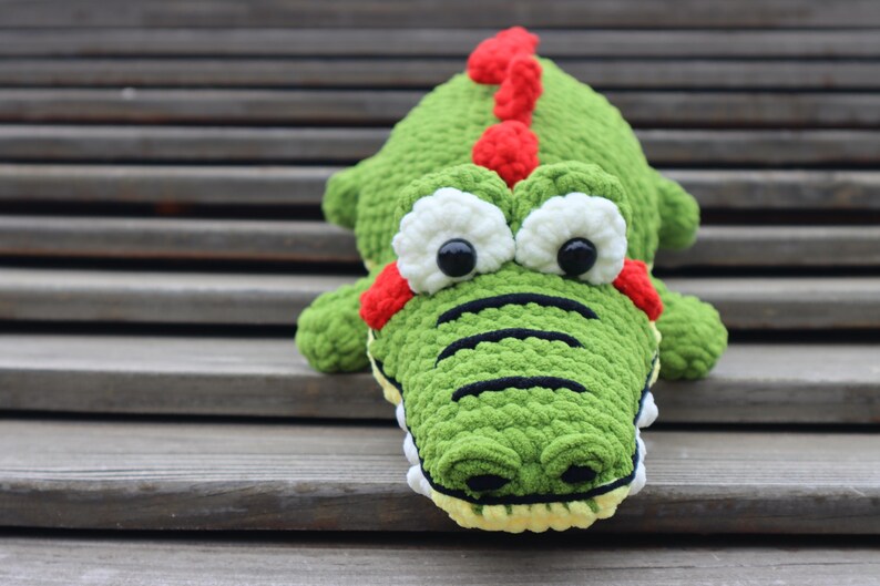K&ouml;nnte beinhalten: Gr&uuml;nes, geh&auml;keltes Alligator-Pl&uuml;schtier mit roten und wei&szlig;en Augen und einem roten, noppigen R&uuml;cken. Der Alligator sitzt auf Holzstufen.