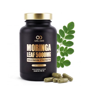 May include: A black bottle with a gold lid labeled "MORINGA LEAF 5000MG MAXIMUM STRENGTH" contains 120 capsules. The bottle is accompanied by green capsules and a sprig of green leaves. The text "VEGAN FRIENDLY CAPSULES MADE IN THE UK" is also visible.