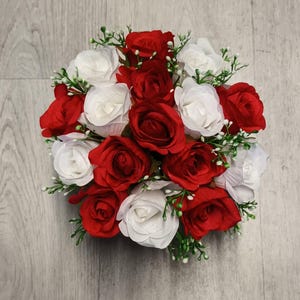 May include: A circular bouquet of red and white roses, interspersed with small green sprigs. The roses are densely packed, forming a full, colourful arrangement. The background is a light grey wooden surface.