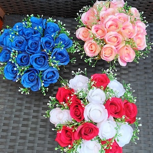 May include: Three bouquets of artificial roses in vibrant colours. One bouquet features deep blue roses, another showcases pink and peach roses, and the third combines red and white roses. Each arrangement includes small white accent flowers and green foliage.
