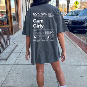 May include: A dark gray t-shirt with the text "100% 100% ORGANIC LEG DAY Gym Girly" printed on the back. The text is in white and has a vintage look. The shirt is being worn by a person.