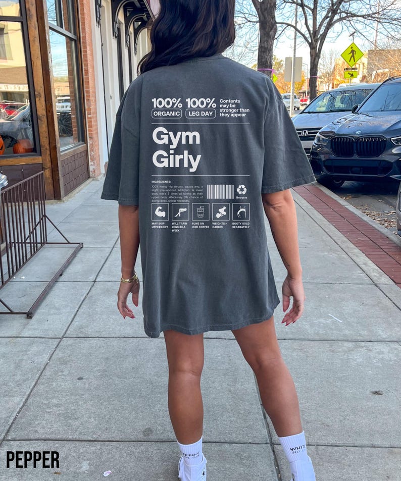 May include: A dark gray t-shirt with the text "100% 100% ORGANIC LEG DAY Gym Girly" printed on the back. The text is in white and has a vintage look. The shirt is being worn by a person.