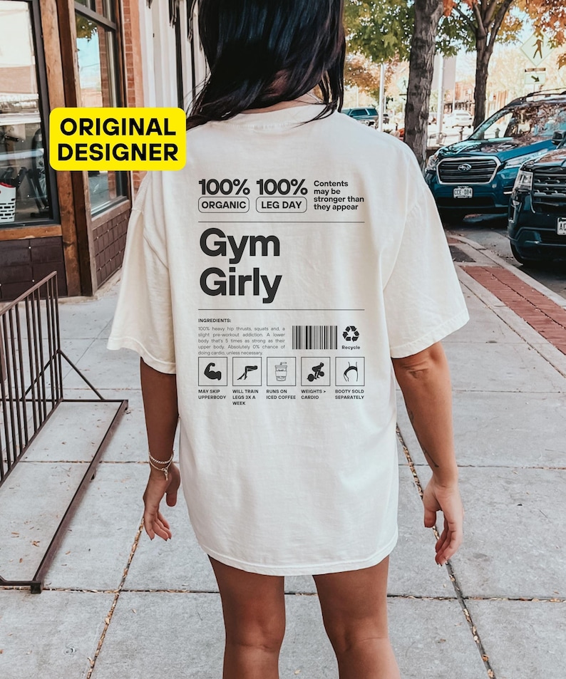May include: White t-shirt with black text that reads "100% 100% Organic Leg Day Gym Girly". The text is in a bold, sans-serif font. Below the text is a black and white graphic that looks like a nutrition label. The label lists ingredients, such as "heavy hip thrusts, squats, and a slight pre-workout addiction", and instructions, such as "May skip upper body a week" and "Will train legs six a week".