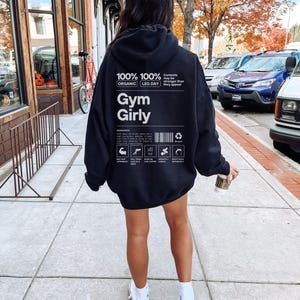 May include: Black hoodie with a white graphic that says "100% 100% Organic Leg Day Gym Girly" and includes a nutrition label with icons. The text "Contents may be stronger than they appear" is also included on the graphic.