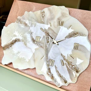 Peut inclure: Une horloge murale artisanale avec un cadran en résine blanc cassé marbré. Des chiffres romains dorés indiquent les heures. La forme irrégulière et les accents dorés incrustés ajoutent à son attrait artistique. Les aiguilles de l'horloge sont dorées.