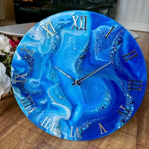 Puede incluir: Un reloj de pared redondo con un diseño veteado azul y blanco. Los números romanos plateados marcan las horas. Las manecillas del reloj son plateadas. El reloj está sobre una superficie de madera.