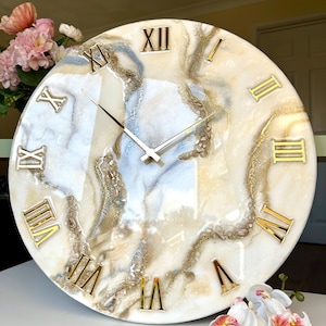 May include: A circular wall clock with a cream-coloured face. It features gold Roman numerals and hands. The clock face has a marbled design in white, beige, and gold. A decorative item for the home.