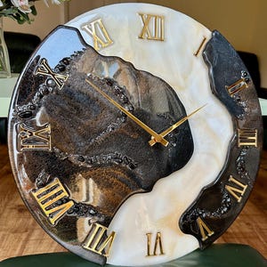 Puede incluir: Reloj de pared redondo con un diseño único. La esfera del reloj presenta una mezcla de resina negra, blanca y dorada con números romanos y manecillas doradas. El diseño abstracto del reloj es adecuado para la decoración del hogar.