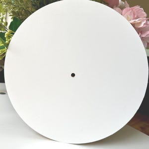 Könnte beinhalten: Ein großes, rundes, weißes Objekt mit einem kleinen, zentrierten schwarzen Loch. Das Objekt ist glatt und scheint aus einem festen Material zu bestehen. Der Hintergrund umfasst Grün und rosa Blumen.