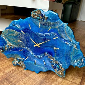 Könnte beinhalten: Eine einzigartige Wanduhr mit blauem und goldenem Harzdesign. Das Zifferblatt hat goldene Zeiger und Markierungen. Dekorative Steine und Kristalle sind in das Harz eingebettet. Die Uhr hat einen Durchmesser von etwa 30 cm.