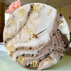 May include: A circular wall clock with a white and brown marbled effect. It has gold Roman numerals and gold hands. Small stones are set in the resin. This decorative clock is a home decor item.