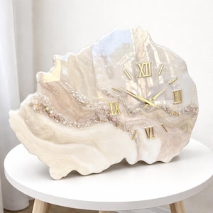 May include: A decorative clock with a unique, organic shape. The clock face features a pearlescent, marbled design with gold Roman numerals and hands. The clock is embellished with small, sparkling crystals and sits on a white table.
