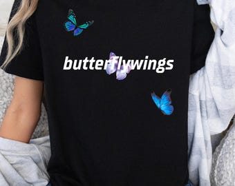 May include: Black t-shirt with the text "butterflywings" in white. The shirt features three butterfly graphics in shades of blue and purple. The butterflies are positioned around the text, creating a visually appealing design.