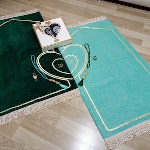 May include: Two prayer mats, one in emerald green and one in teal, with gold trim and fringing. A white box with a heart design and a gold ribbon sits between the mats. Prayer beads and a watch are also visible.