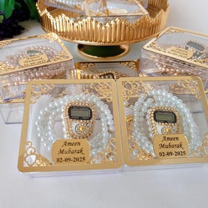 Puede incluir: Dos cajas de plástico transparente con bordes dorados, cada una contiene un mala de oración de cuentas blancas con un contador digital y una pluma blanca. Las cajas tienen una inscripción dorada que dice "Ameen Mubarak 02-09-2025".