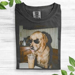 May include: A charcoal gray crew neck t-shirt featuring a graphic print of a golden retriever wearing black sunglasses and drinking from a clear plastic cup with a straw. The shirt has a Comfort Colors brand label on the inside of the collar.