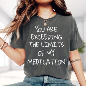 May include: A charcoal gray short-sleeve t-shirt featuring the white printed text YOU ARE EXCEEDING THE LIMITS OF MY MEDICATION. The shirt has a crew neckline and a relaxed fit, constructed from a soft, washed-out fabric with a vintage appearance.