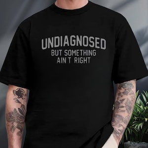 May include: A black short-sleeved t-shirt featuring the text "UNDIAGNOSED" in a large, arched, grey sans-serif font, followed by the text "BUT SOMETHING AIN'T RIGHT" in a smaller, straight, grey sans-serif font centered on the chest area.
