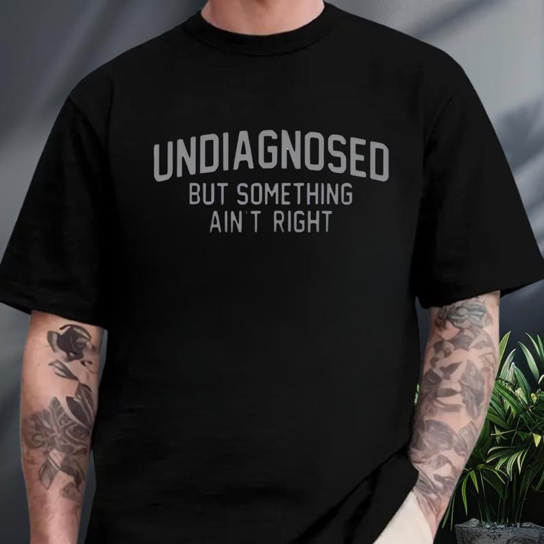 May include: A black short-sleeved t-shirt featuring the text "UNDIAGNOSED" in a large, arched, grey sans-serif font, followed by the text "BUT SOMETHING AIN'T RIGHT" in a smaller, straight, grey sans-serif font centered on the chest area.