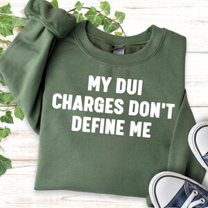 May include: An olive green crewneck sweatshirt featuring the white text "MY DUI CHARGES DON'T DEFINE ME" printed in a bold, sans-serif font across the chest. The garment has long sleeves with ribbed cuffs and a ribbed neckline.