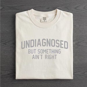May include: A cream-colored short-sleeve t-shirt with a crew neckline. The shirt features the gray text "UNDIAGNOSED BUT SOMETHING AIN'T RIGHT" printed in a bold, sans-serif font across the chest. A Comfort Colors brand label is visible at the neck.