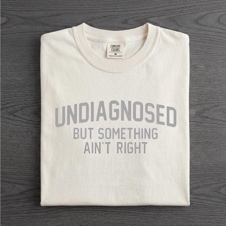 May include: A cream-colored short-sleeve t-shirt with a crew neckline. The shirt features the gray text "UNDIAGNOSED BUT SOMETHING AIN'T RIGHT" printed in a bold, sans-serif font across the chest. A Comfort Colors brand label is visible at the neck.