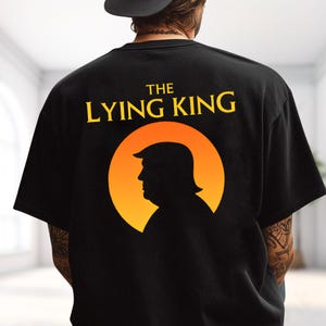 May include: A black short-sleeved t-shirt featuring a graphic on the back. The design includes the yellow text THE LYING KING above a circular orange and yellow gradient sun icon containing the black silhouette profile of a person.