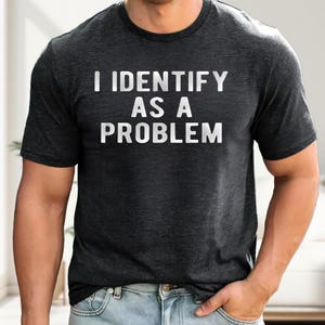 May include: A charcoal gray short-sleeved t-shirt featuring the white text "I IDENTIFY AS A PROBLEM" printed in a bold, sans-serif font across the chest. The garment has a crew neckline and a heathered fabric texture throughout the material.