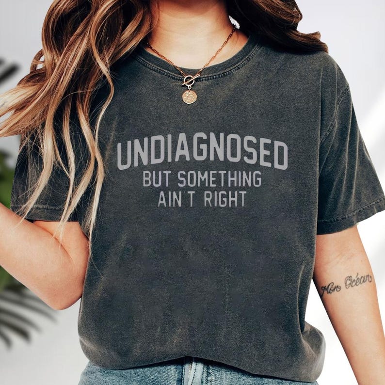 May include: A charcoal gray short-sleeve t-shirt with a crew neckline. The shirt features the text "UNDIAGNOSED BUT SOMETHING AIN'T RIGHT" printed in a light gray, sans-serif font across the center of the chest area on the front of the garment.