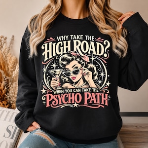 May include: A black long-sleeve sweatshirt featuring a graphic illustration of a person with vintage-style hair, sunglasses, and a cocktail glass. The text reads, "WHY TAKE THE HIGH ROAD? WHEN YOU CAN TAKE THE PSYCHO PATH" in cream and pink lettering.