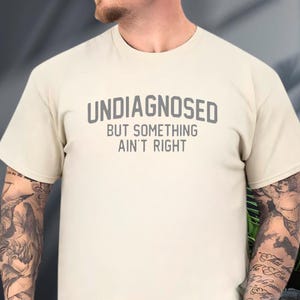 May include: A cream-colored short-sleeve t-shirt featuring the dark gray text "UNDIAGNOSED BUT SOMETHING AIN'T RIGHT" printed in a bold, sans-serif font across the chest. The garment has a classic crew neckline and a standard, relaxed fit.