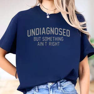 May include: A navy blue short-sleeved t-shirt featuring the text "UNDIAGNOSED BUT SOMETHING AIN'T RIGHT" printed in a light gray, sans-serif font across the chest. The shirt has a crew neckline and a standard fit, paired with blue denim jeans.