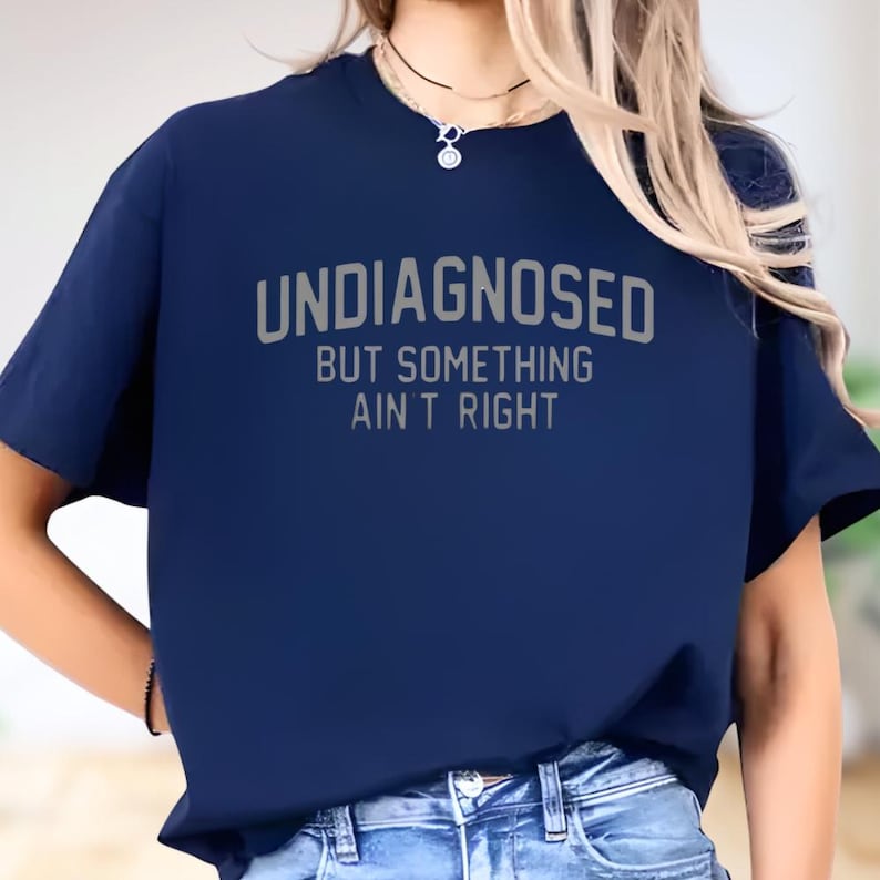 May include: A navy blue short-sleeved t-shirt featuring the text "UNDIAGNOSED BUT SOMETHING AIN'T RIGHT" printed in a light gray, sans-serif font across the chest. The shirt has a crew neckline and a standard fit, paired with blue denim jeans.