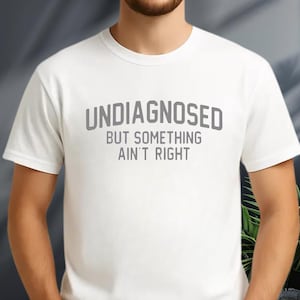 May include: A white short-sleeved t-shirt featuring the grey text "UNDIAGNOSED BUT SOMETHING AIN'T RIGHT" printed in a bold, sans-serif font across the chest. The garment has a classic crew neckline and a standard fit, providing a clean look.