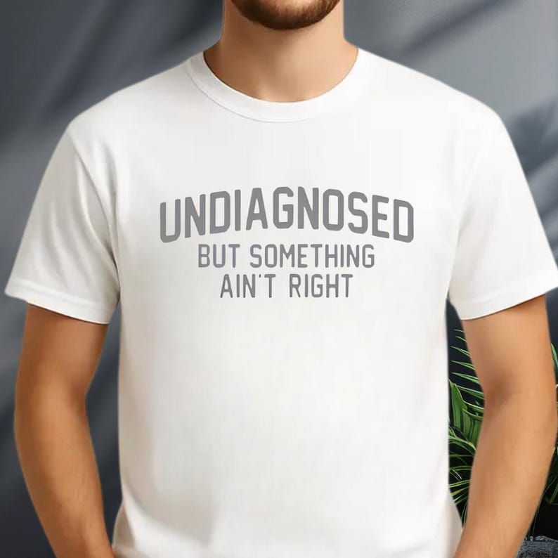 May include: A white short-sleeved t-shirt featuring the grey text "UNDIAGNOSED BUT SOMETHING AIN'T RIGHT" printed in a bold, sans-serif font across the chest. The garment has a classic crew neckline and a standard fit, providing a clean look.