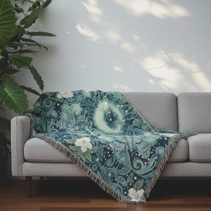 Puede incluir: Una manta tejida con un diseño botánico y celestial detallado. La manta presenta una luna creciente, estrellas, flores y follaje en tonos de verde azulado, azul marino y blanco. La manta está sobre un sofá gris.