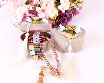 May include: A set of prayer beads with a digital counter, a tassel, and a small box with a floral design. The beads are a light brown color and the box is a light beige color.